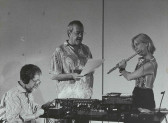Black-and-white photograph of three figures performing together. On the left, a figure in a striped shirt and glasses sits down in front of a table full of audio equipment, wearing headphones. In the middle, a figure in a graphic shirt stands and reads off a piece of paper. The rightmost figure wears a glitter and mesh top and plays the flute standing.
