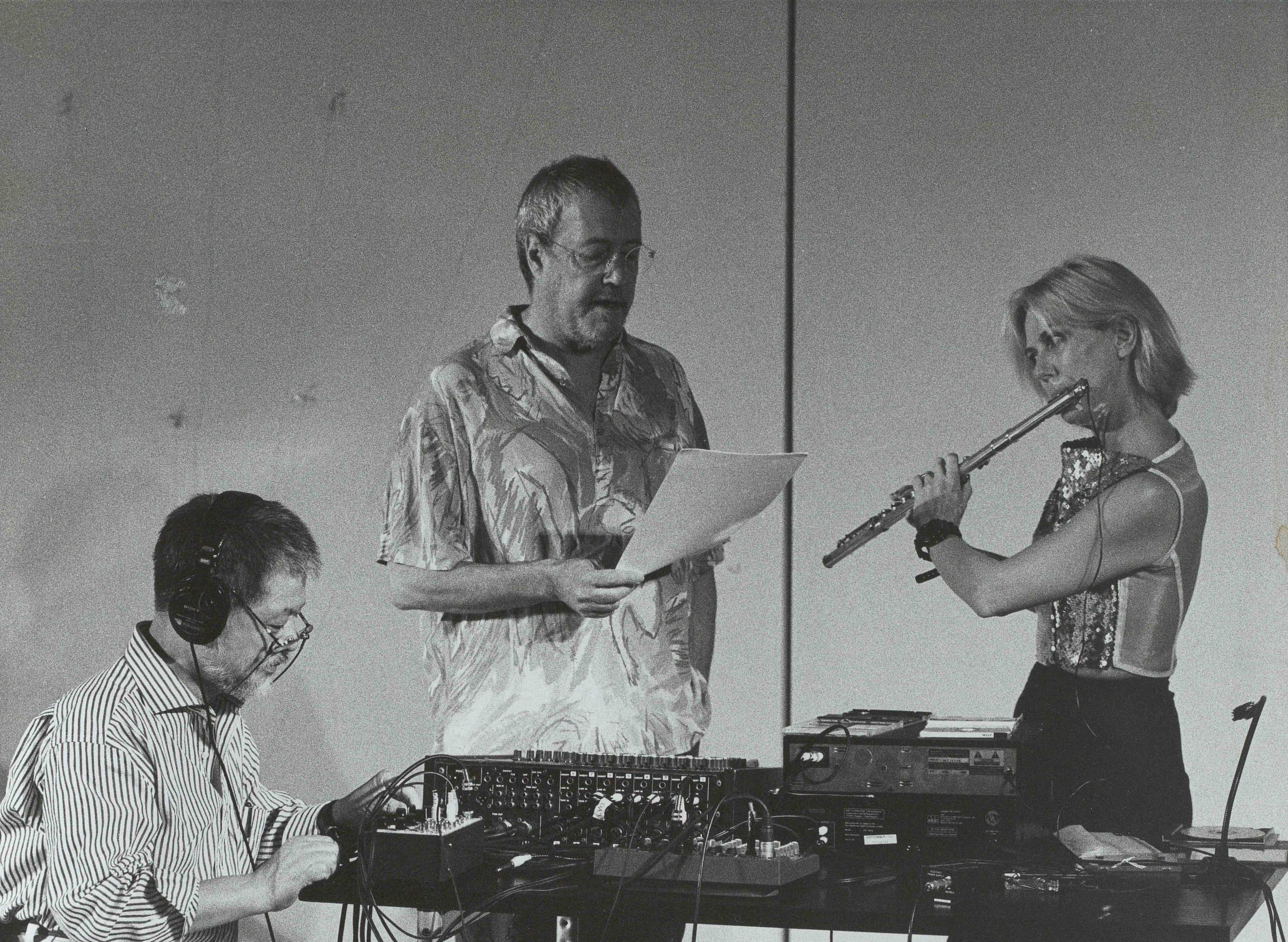 Black-and-white photograph of three figures performing together. On the left, a figure in a striped shirt and glasses sits down in front of a table full of audio equipment, wearing headphones. In the middle, a figure in a graphic shirt stands and reads off a piece of paper. The rightmost figure wears a glitter and mesh top and plays the flute standing.