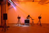A color photograph of a figure standing behind audio mixing equipment and a microphone in front of an orange background. A white light shines down on them from the left.