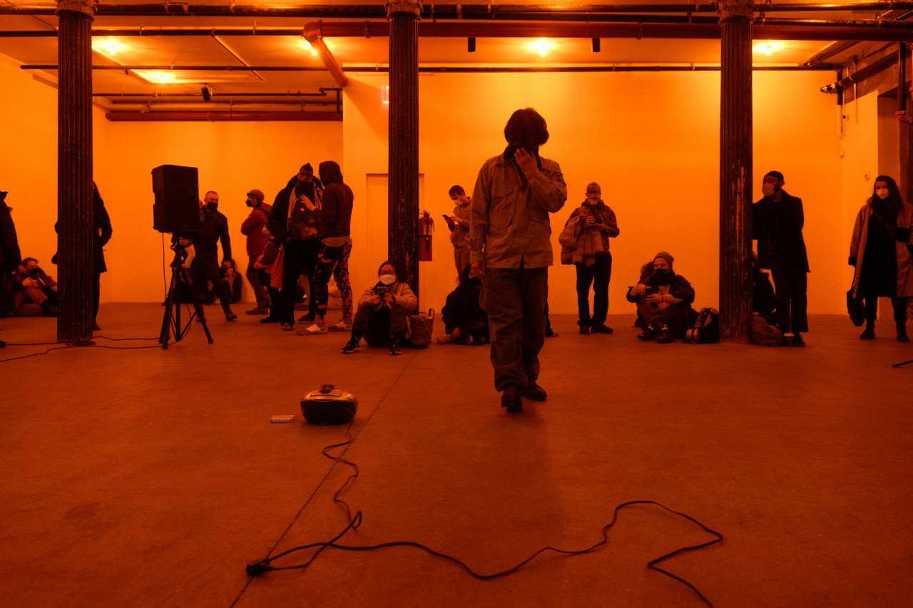 A color photograph of a figure standing in an orange-lit space next to a small piece of audio equipment with a wire on the floor. A crowd looks on in the background.