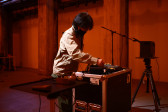 A color photograph of a figure in an orange and white-lit space, leaning forward slightly to adjust a dial on a piece of audio equipment.
