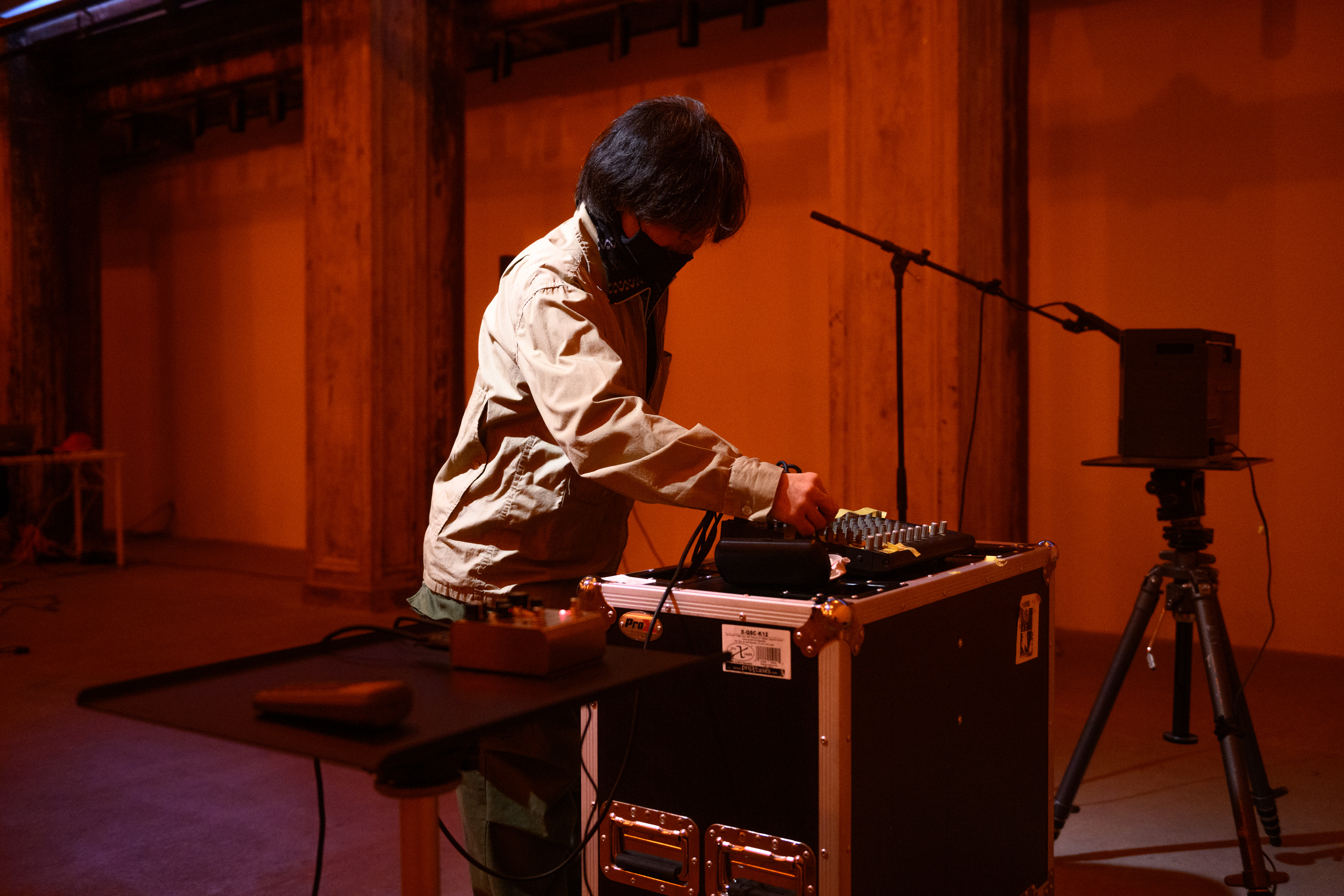 A color photograph of a figure in an orange and white-lit space, leaning forward slightly to adjust a dial on a piece of audio equipment.