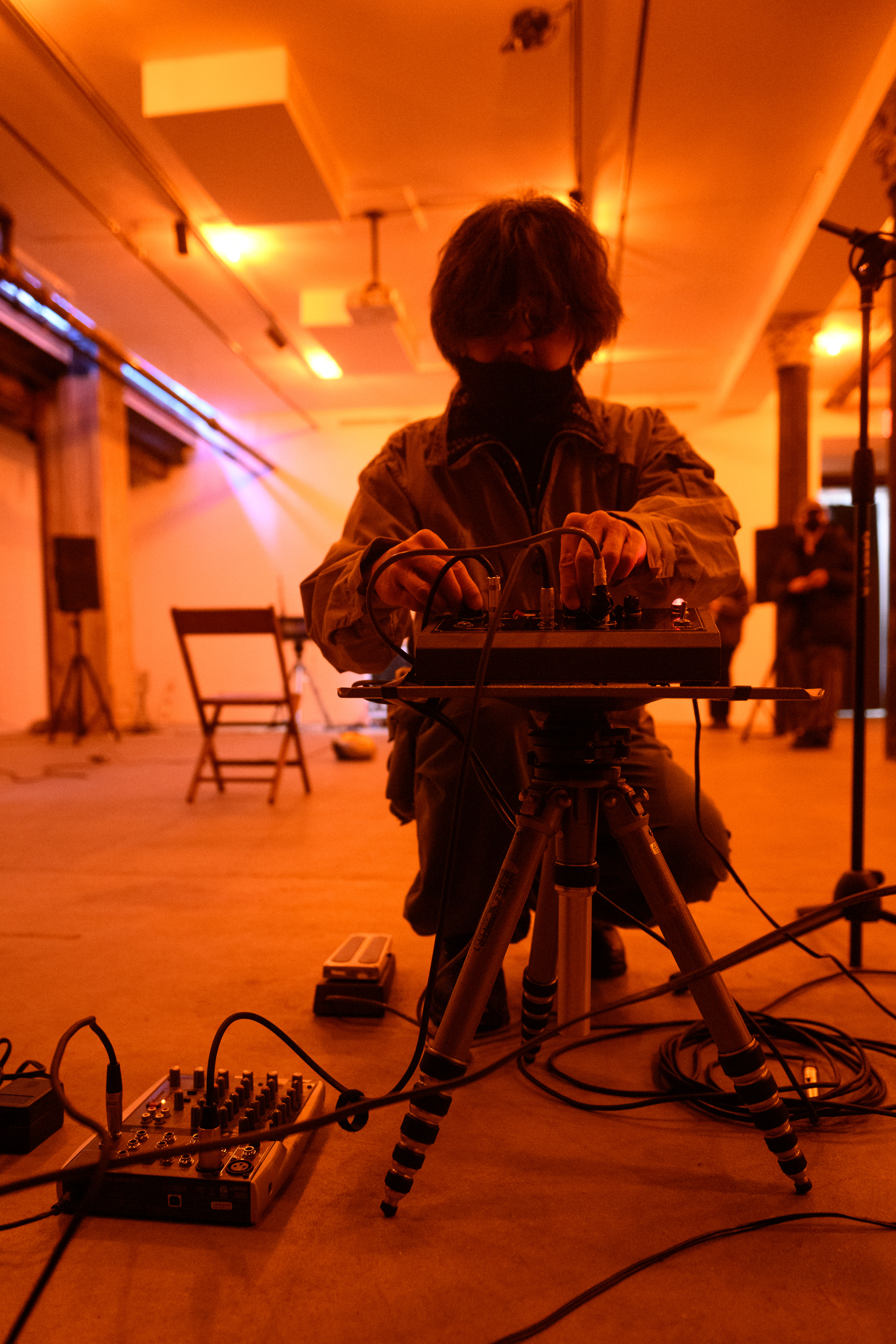 A color photograph of a figure standing in an orange-lit space filled with various audio equipment and a stereo player. They crouch down to press adjust a dial on an audio mixer.