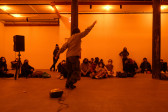 A color photograph of a figure with raised arms and in motion in an orange-lit space next to a small piece of audio equipment with a wire on the floor. A crowd looks on in the background.