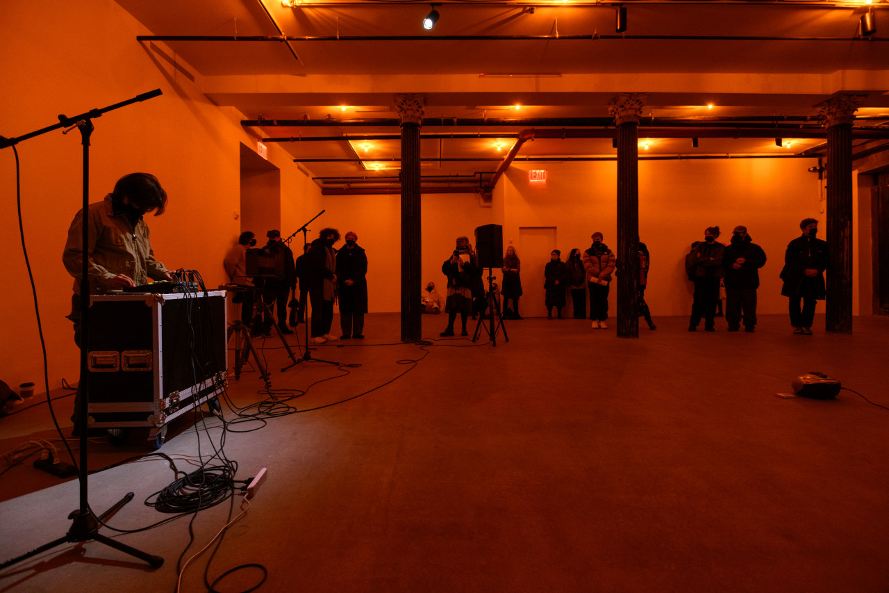 A color photograph of a figure standing behind audio mixing equipment and a microphone in front of an orange background. A crowd looks on.