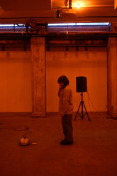 A color photograph of a figure standing in an orange-lit space next to a small piece of audio equipment with a wire on the floor. A speaker is visible in the background.