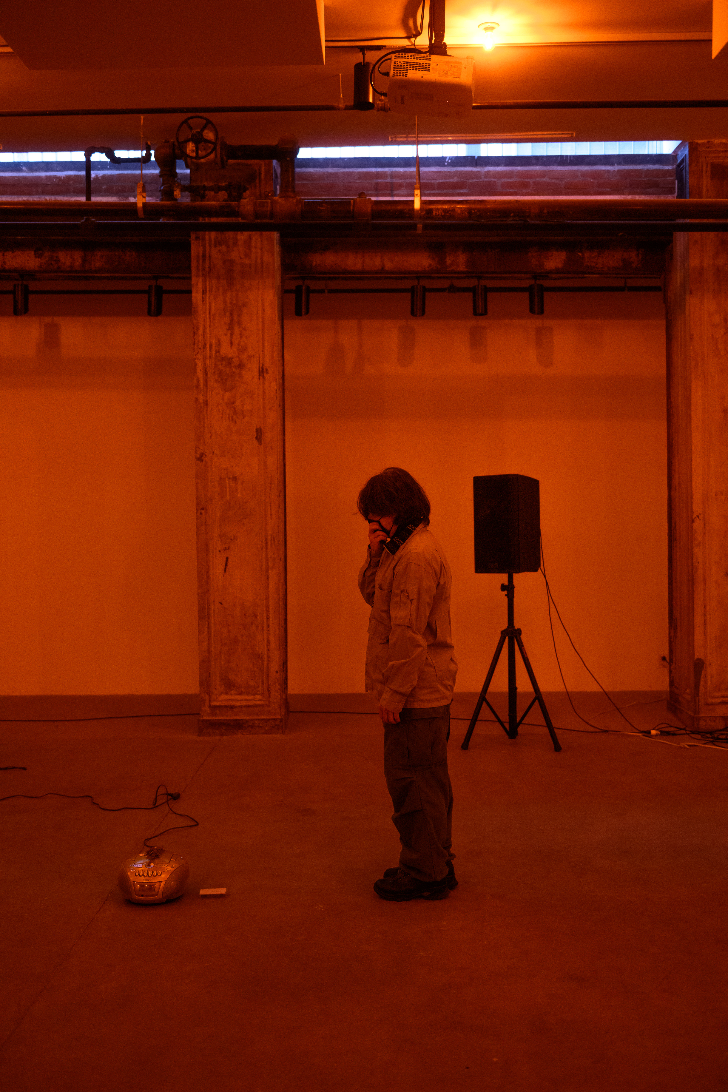 A color photograph of a figure standing in an orange-lit space next to a small piece of audio equipment with a wire on the floor. A speaker is visible in the background.