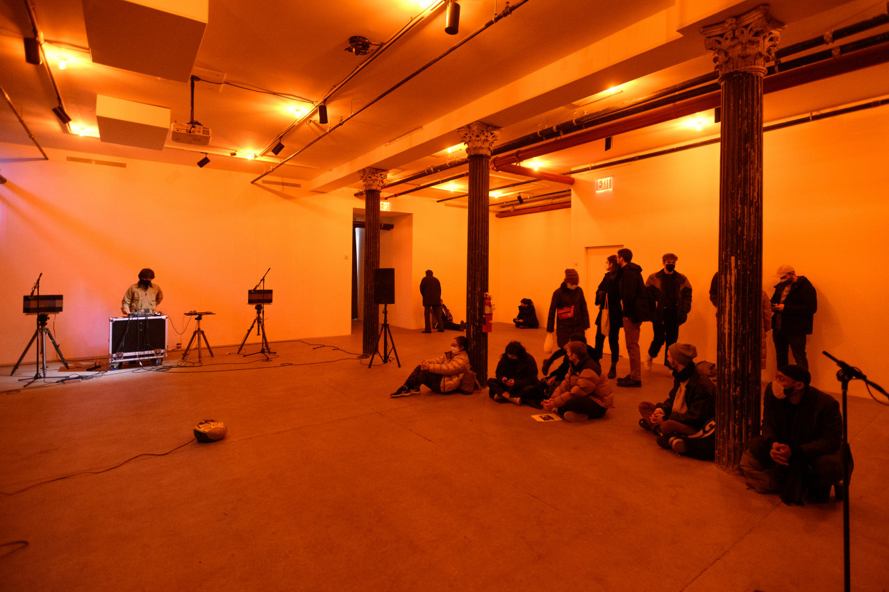 A color photograph of a figure standing behind audio mixing equipment and a microphone in an orange-lit space. A crowd looks on.