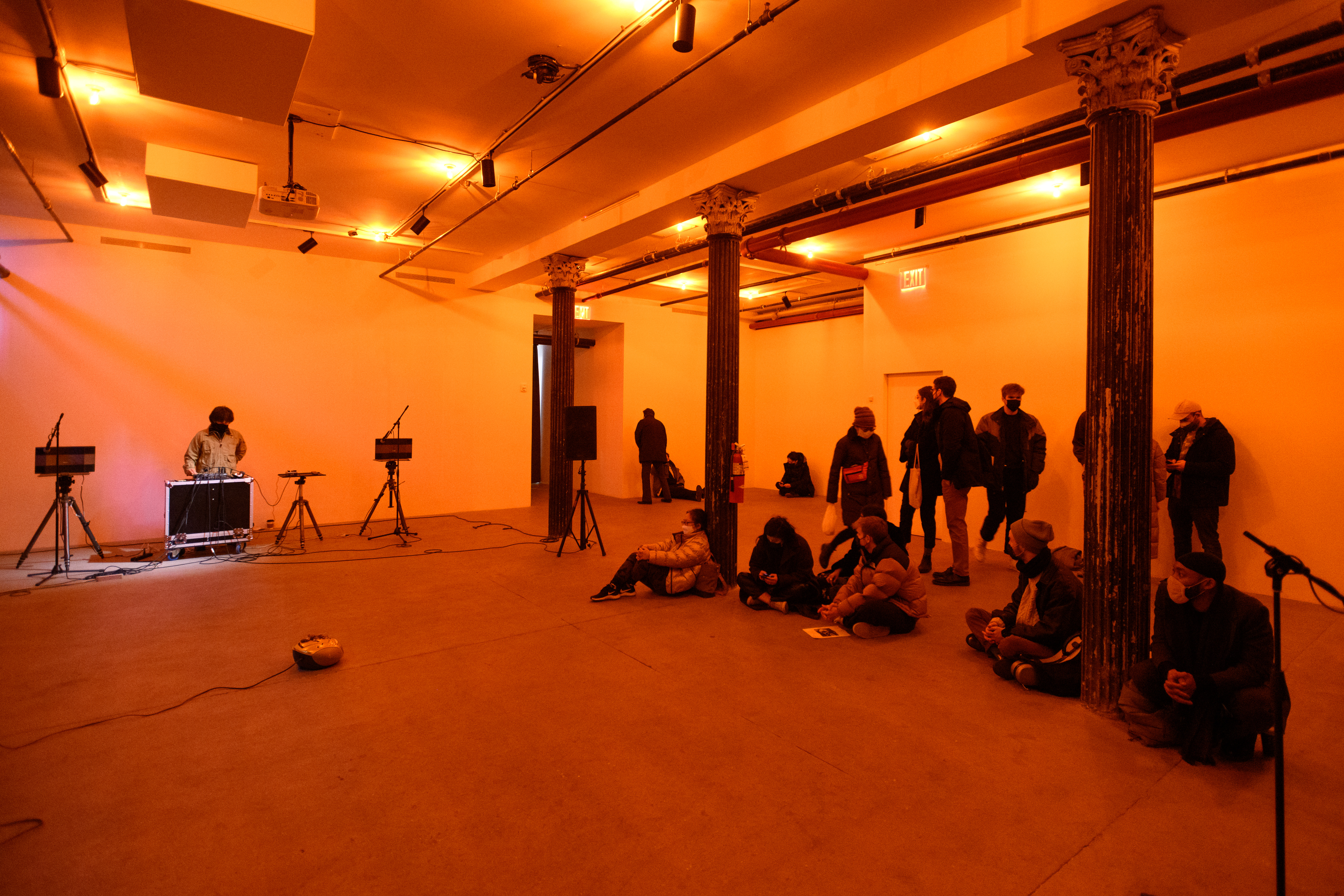 A color photograph of a figure standing behind audio mixing equipment and a microphone in an orange-lit space. A crowd looks on.