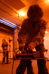 A color photograph of a figure standing in an orange-lit space filled with various audio equipment. They lean down to press adjust a dial on an audio mixer.