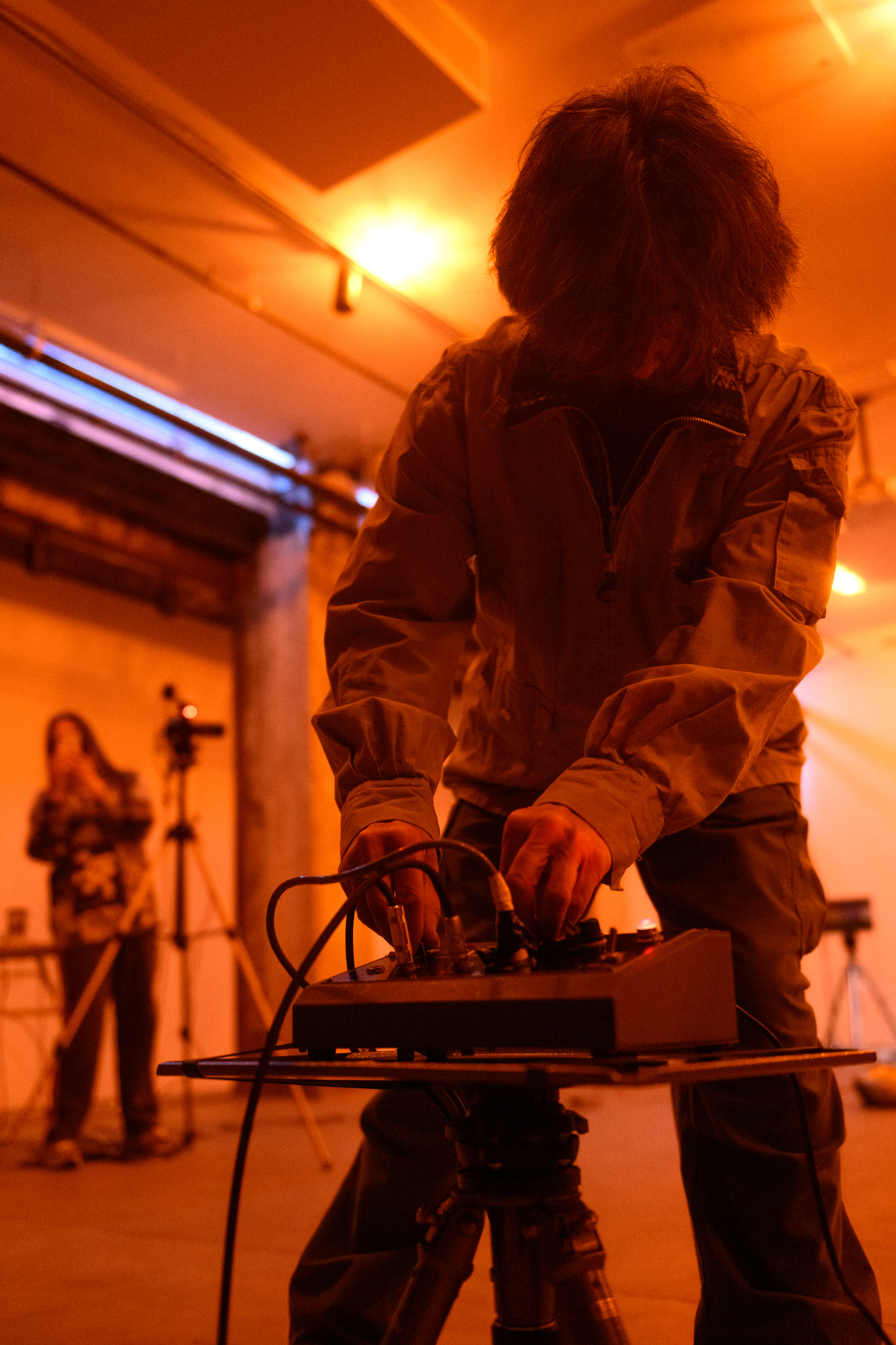 A color photograph of a figure standing in an orange-lit space filled with various audio equipment. They lean down to press adjust a dial on an audio mixer.
