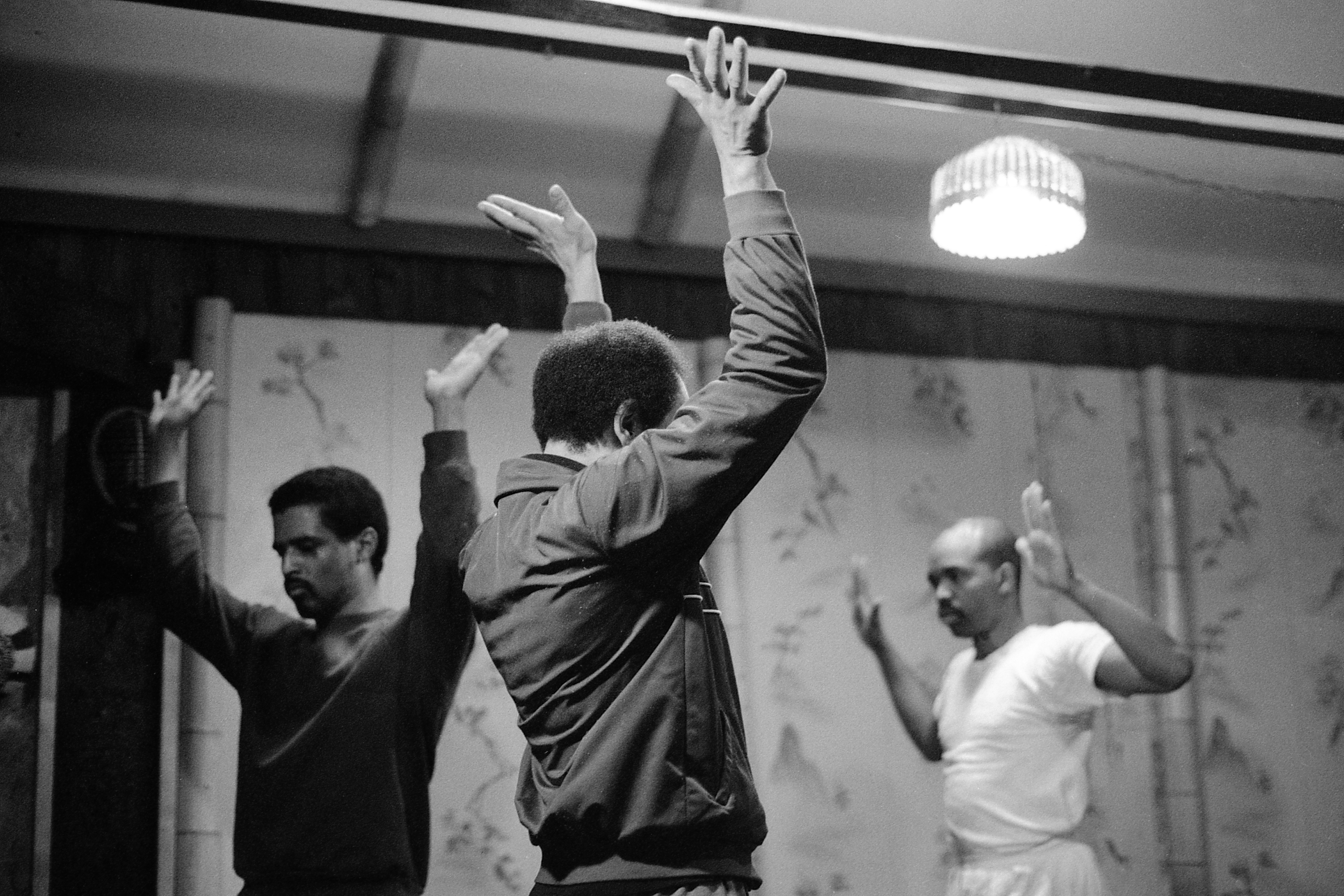 A black-and-white photograph depicting three men equally distanced from one another. They hold their hands up to the ceiling with their palms out and stand near a floral printed wall.