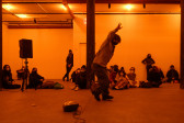 A color photograph of a figure with raised arms and in motion in an orange-lit space next to a small piece of audio equipment with a wire on the floor. A crowd looks on in the background.