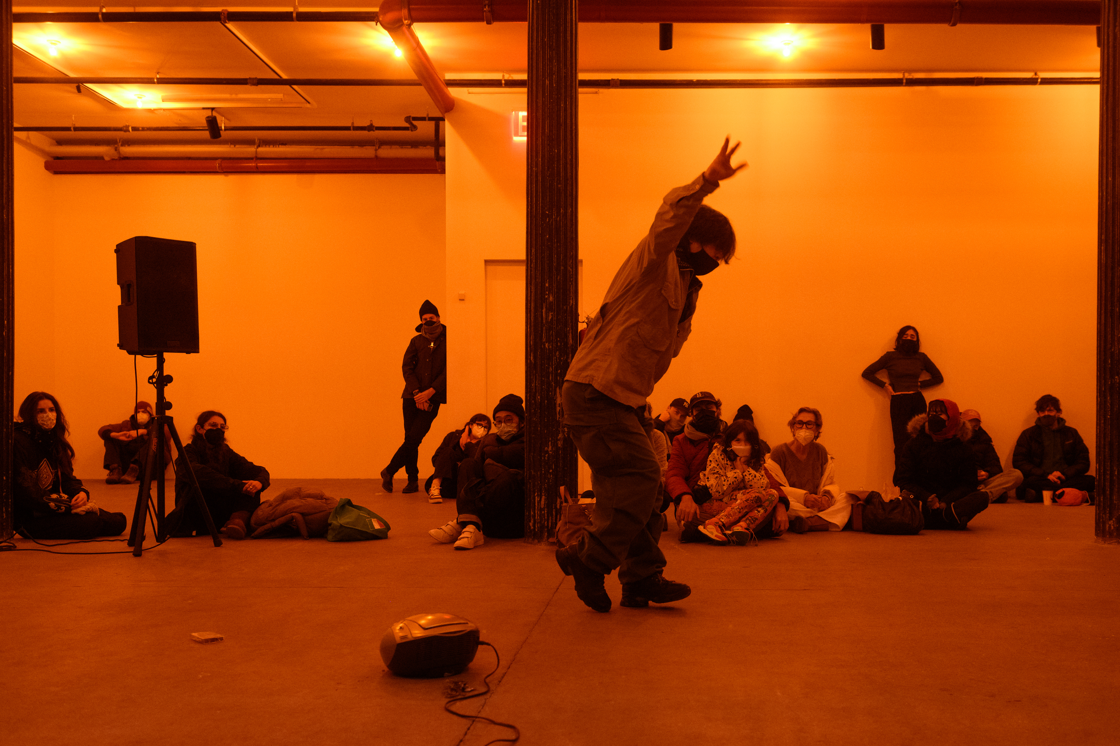 A color photograph of a figure with raised arms and in motion in an orange-lit space next to a small piece of audio equipment with a wire on the floor. A crowd looks on in the background.