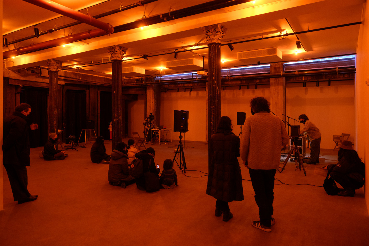 A color photograph of a figure standing behind audio mixing equipment and a microphone in an orange-lit space. A crowd looks on.
