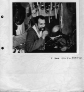 Image of a photograph attached to a piece of paper. The photograph is of a seated person with a mustache, swifly waving their hands up at chest height. A mirror on the left reflects the figure of a standing person in white pants and a dark shirt.