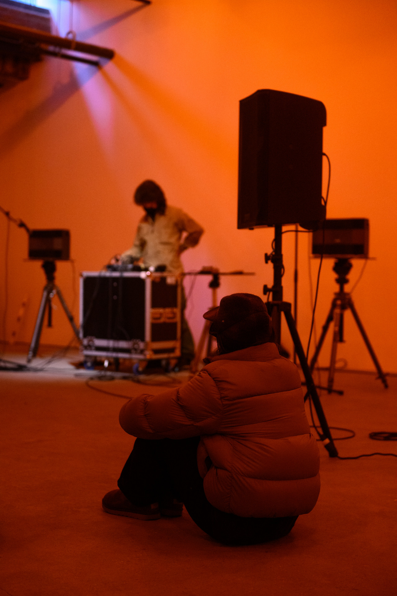 A color photograph of a figure standing behind audio mixing equipment and a microphone in an orange-lit space. A seated observer watches in the foreground.