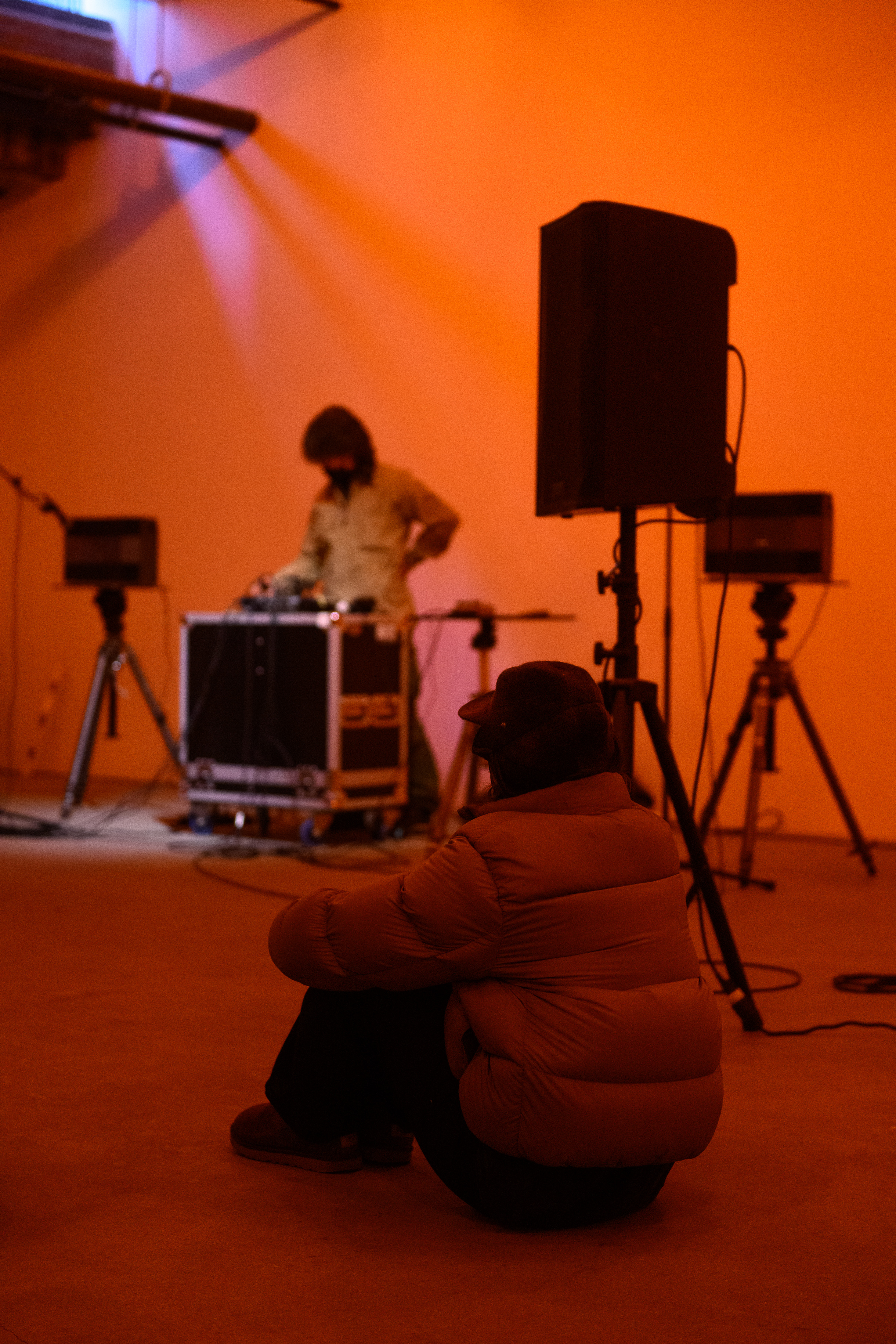 A color photograph of a figure standing behind audio mixing equipment and a microphone in an orange-lit space. A seated observer watches in the foreground.