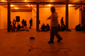 A color photograph of a figure in motion in an orange-lit space next to a small piece of audio equipment with a wire on the floor. A crowd looks on in the background.
