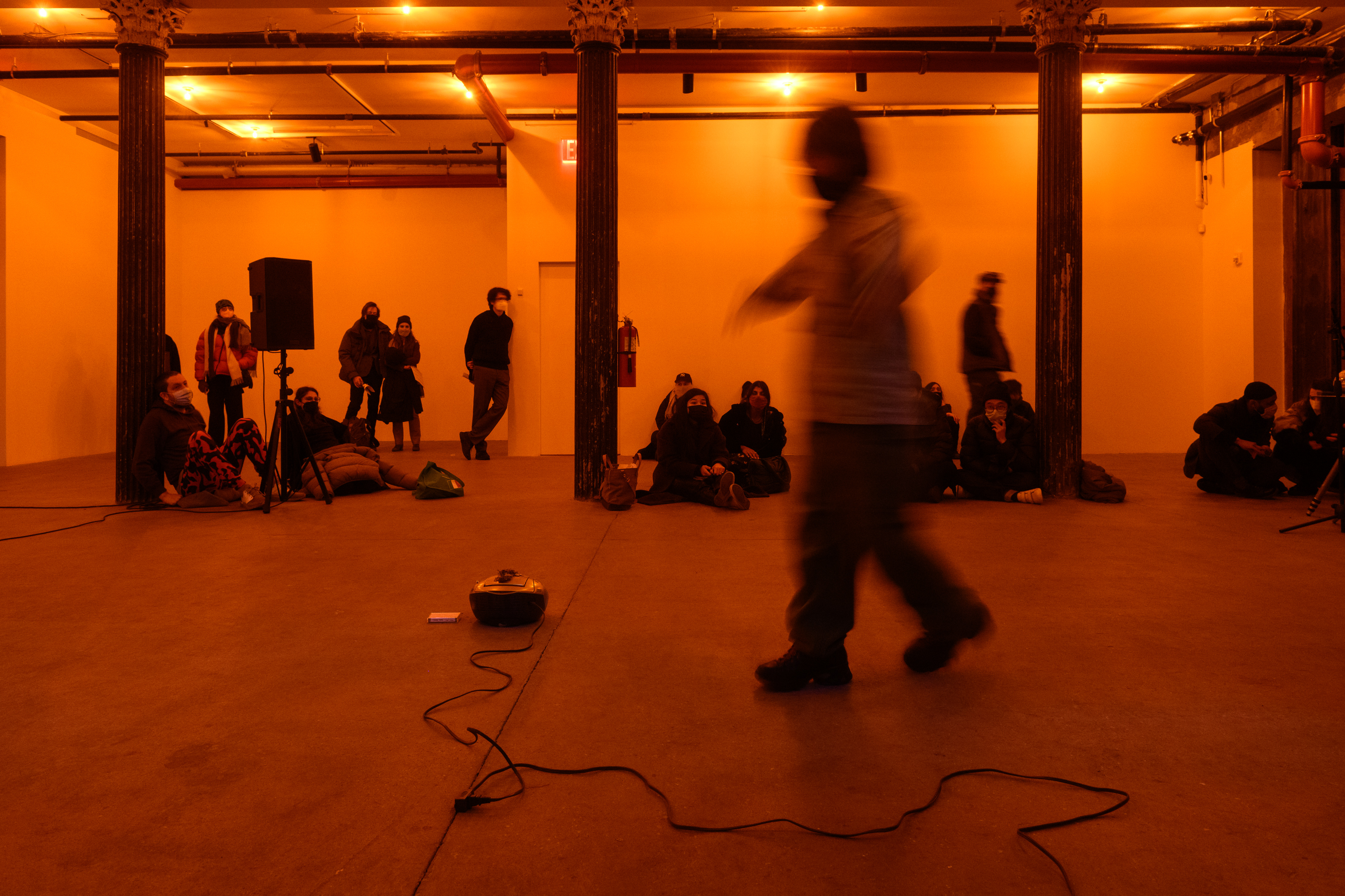 A color photograph of a figure in motion in an orange-lit space next to a small piece of audio equipment with a wire on the floor. A crowd looks on in the background.