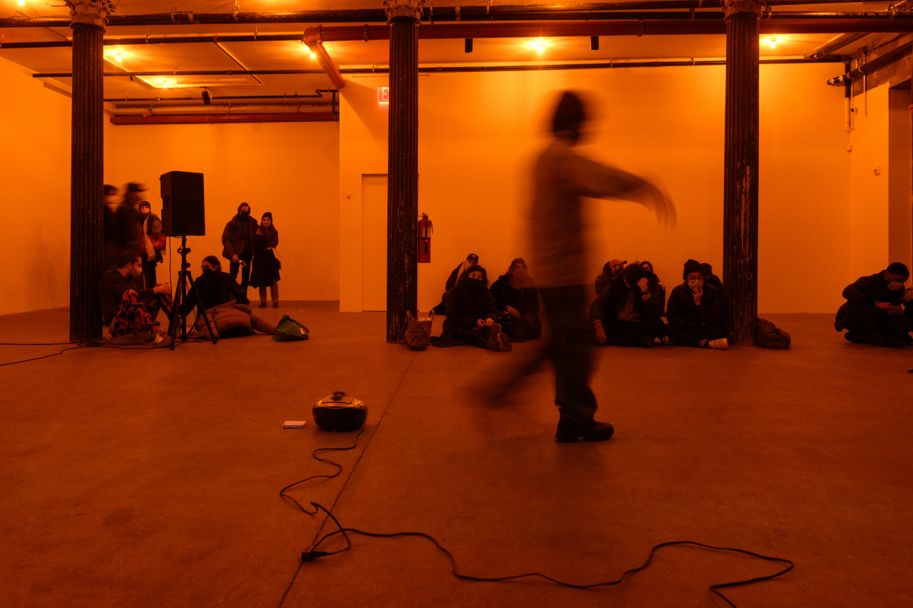 A color photograph of a figure in motion in an orange-lit space next to a small piece of audio equipment with a wire on the floor. A crowd looks on in the background.