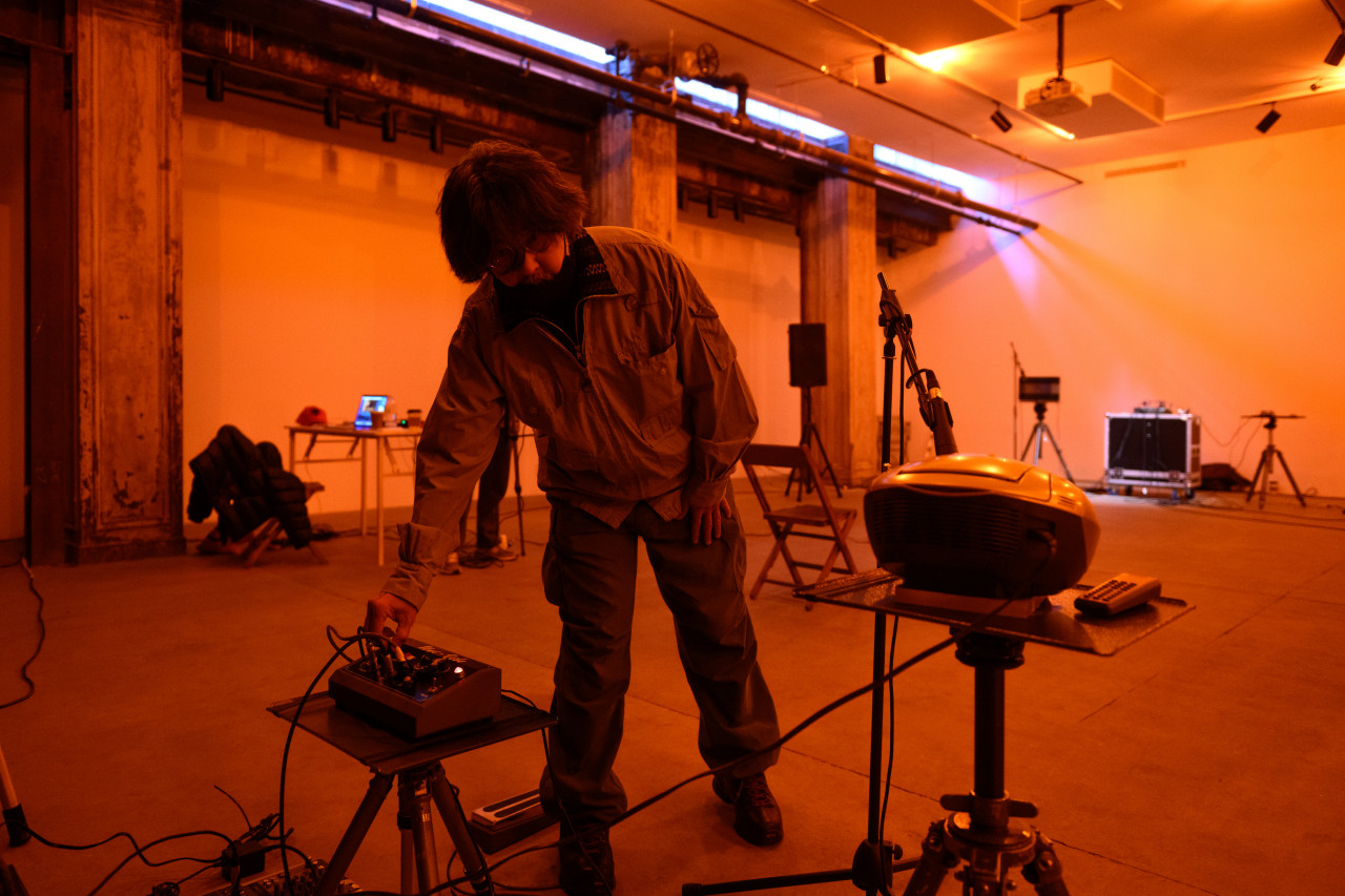 A color photograph of a figure standing in an orange-lit space filled with various audio equipment and a stereo player. They lean down to press adjust a dial on an audio mixer.