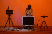 A color photograph of a figure standing behind audio mixing equipment and a microphone in front of an orange background.