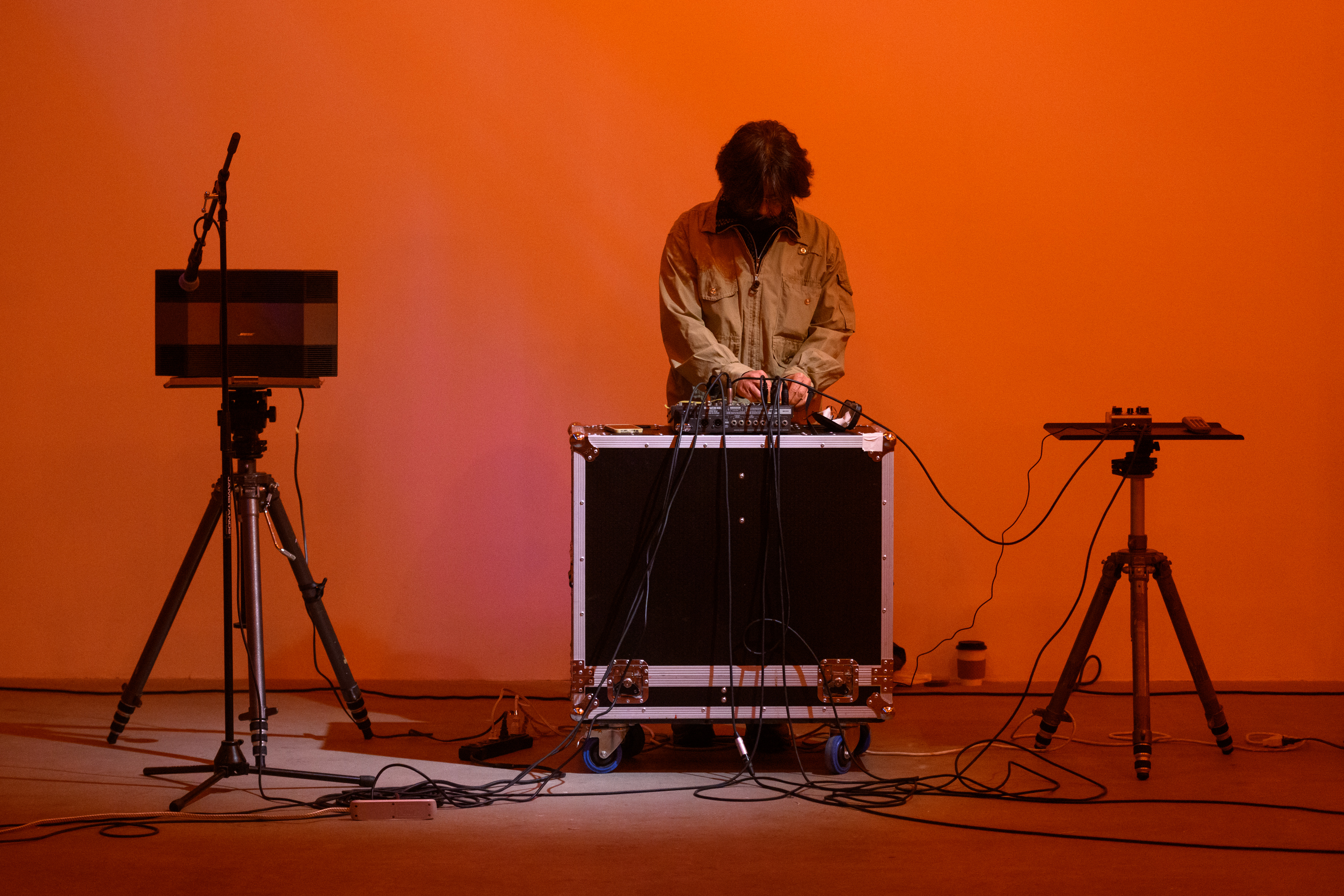 A color photograph of a figure standing behind audio mixing equipment and a microphone in front of an orange background.