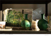 Photograph of a series of objects clustered on a dusty shelf, illuminated by soft daylight. Two identical green vases and two clear cups sit on the right side of the shelf. A photograph of several tomato plant stalks and other flowers is causally propped behind the leftmost vase. Stacks of paper, a plastic bag, and used candles are scattered behind these objects. Small text at the lower left and right corners of the image reads, 