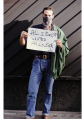 Photograph of a youthful figure with short brown hair wearing blue jeans and a dark shirt with a green jacket tossed over their left shoulder. A black bandana covers their mouth, obscuring the lower half of their face. They hold a sign in front of their chest with both hands. The phrase, 