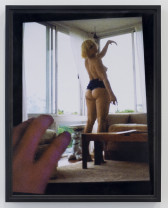 A framed photo of a hand in the lower left corner adjusting a skewed image of K8 Hardy partially undressed in a windowed room, looking over her shoulder with her left arm held aloft.