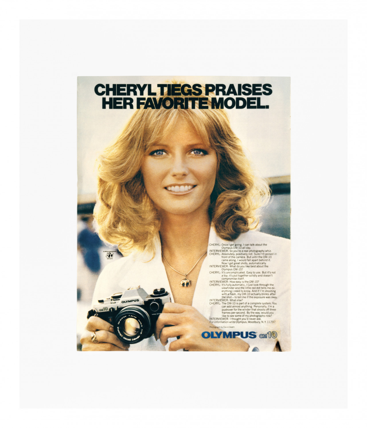 A color photo advertisement for an Olympus OM-10 camera, depicting a woman holding the camera and smiling, looking just past the viewer. The image is titled "CHERYL TIEGS PRAISES HER FAVORITE MODEL." A short interview is printed at the bottom right of the image, where Cheryl goes back and forth with an "interviewer" about her love of the camera.