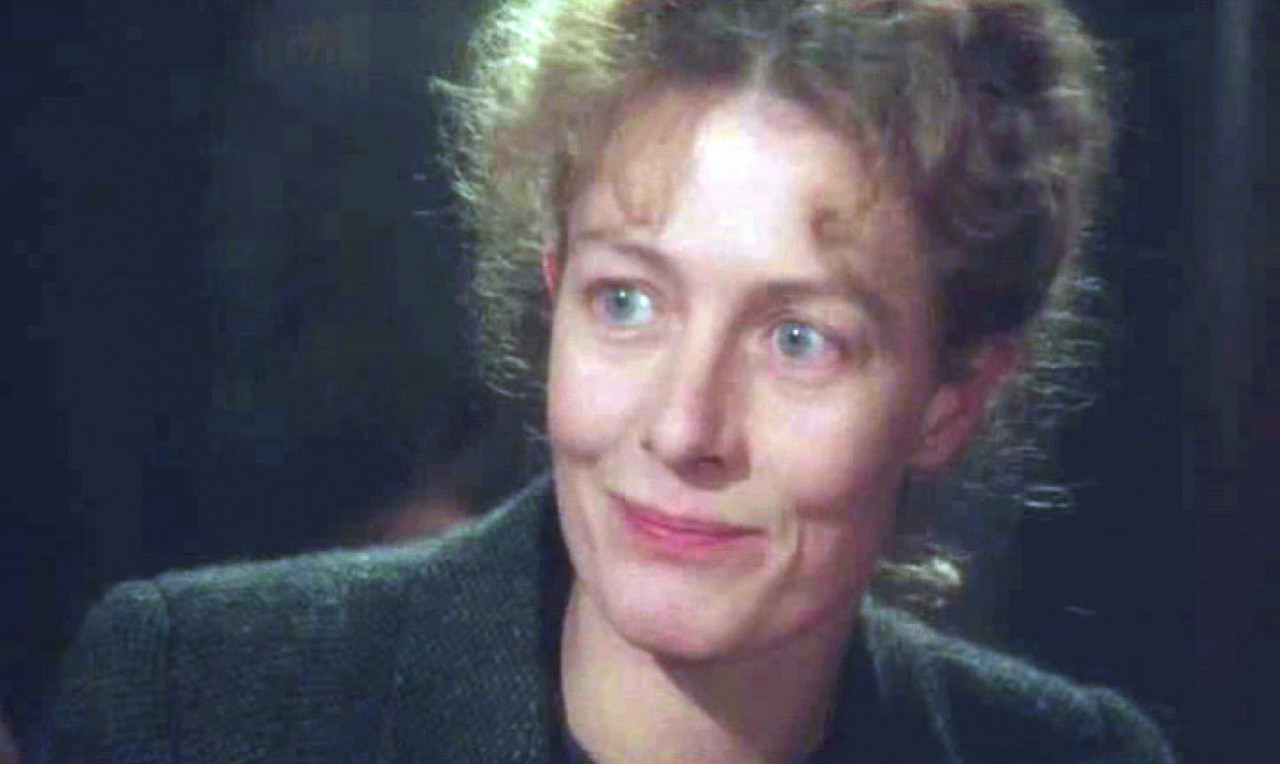 An image of a woman with frizzy hair, smiling and looking past the viewer. The image is slightly distorted, as if from a VHS tape.