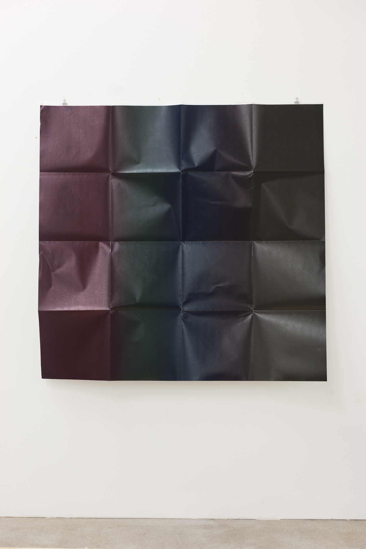 A large, square, sheet of paper, which has been folded into a 4x4 grid, unfolded, and hung on a white wall, leaving the creases. It is covered in a four-color gradient, from dark red, green, blue, to black, left to right, roughly not overlapping with the creases.