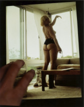 A framed photo of a hand in the lower left corner adjusting a skewed image of K8 Hardy partially undressed in a windowed room, looking over her shoulder with her left arm held aloft.