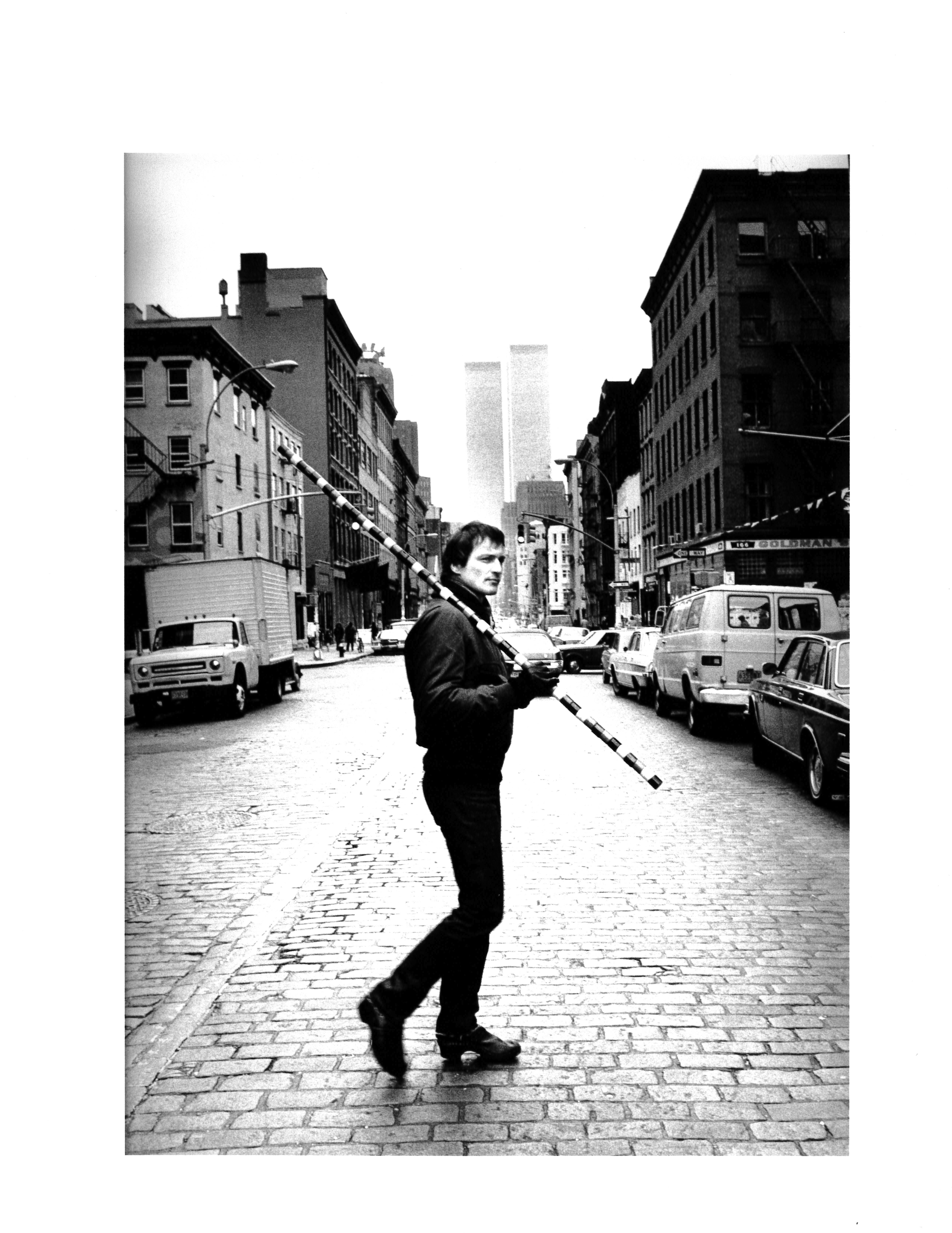 Black and white photo of Andre Cadere holding a striped stick over one shoulder and crossing the street in New York City.