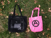 Two Artists Space tote bags lay in the autumn grass. One is black with a white line drawing, the other is pink with a black bold graphic.