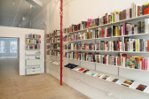 A photo of the bookstore at 55 Walker Street - the front half is a bookstore, while the back half is an empty exhibition/performance space. Both have white walls and a light wood floor. They are divided by a temporary wall with a large doorway, which leaves a large gap between the top of the temporary wall and the ceiling. The back half is empty, with windows letting in light on the furthest wall. The front half has bookshelves adorning the walls, with a lower shelf to keep some books on display. A red pipe runs floor-to-ceiling between two columns of shelves.