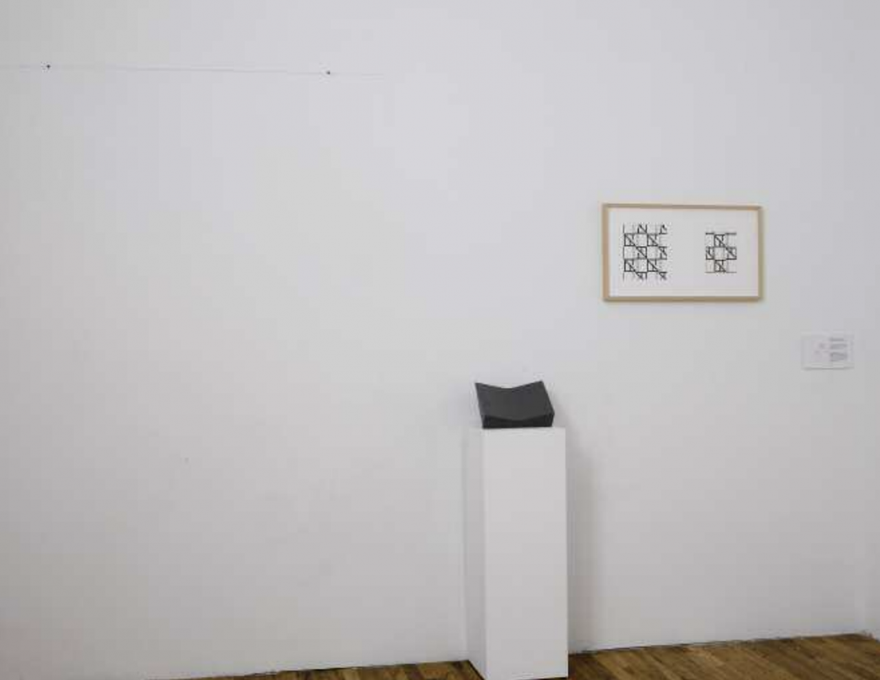 The photograph shows a framed image with black geometric patterns mounted on a white wall with a black sculpture to its left sitting on a white plinth.