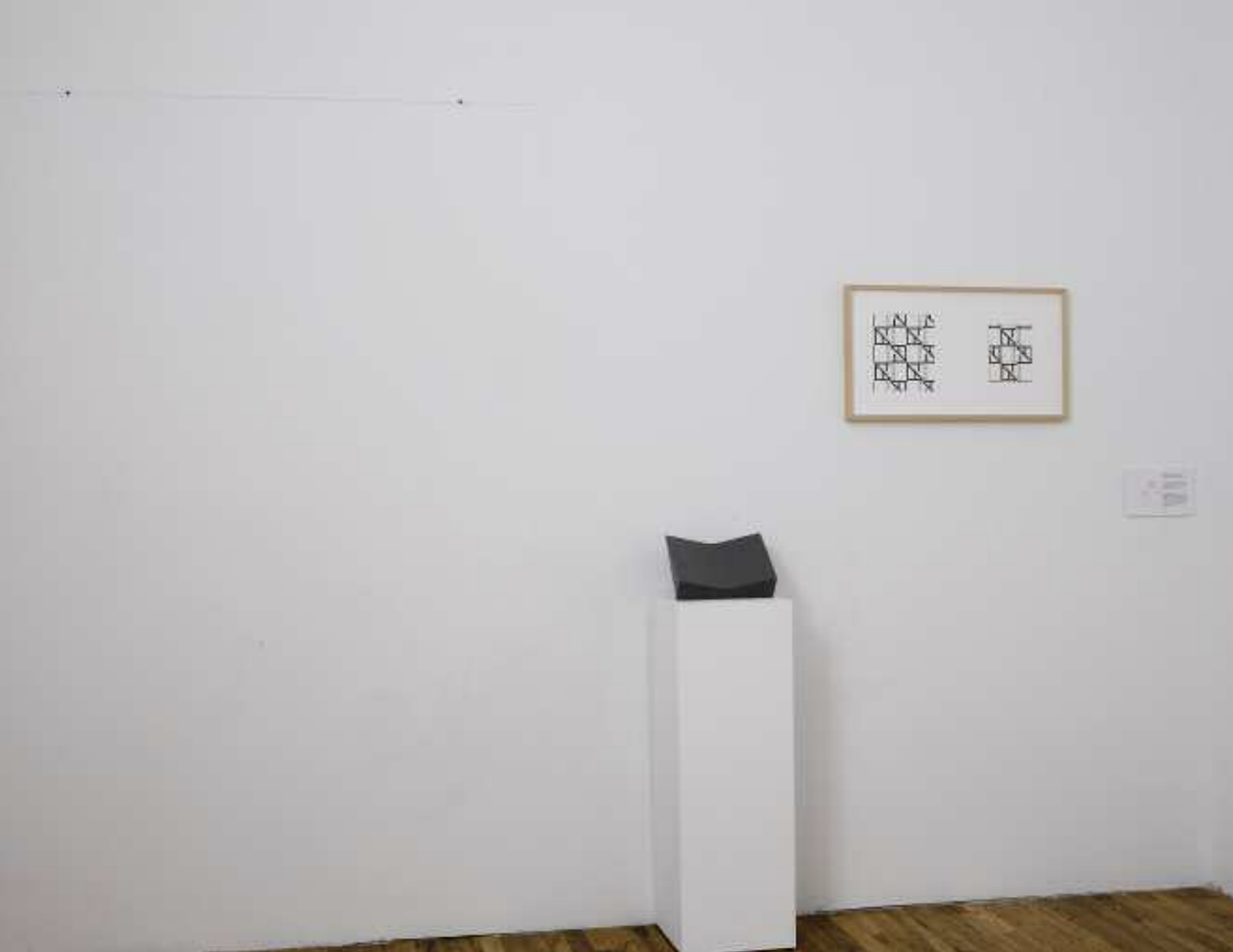 The photograph shows a framed image with black geometric patterns mounted on a white wall with a black sculpture to its left sitting on a white plinth.