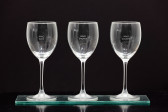 Three wine glasses sit next to each other on a glass shelf with a black background. Each glass has text lasered into the front. From left to right the first says, 
