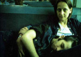 Two people sit with a dark blue seat back behind them. One, on the right of the image, looks to the camera with the other lays asleep on their lap across the bottom of the image.