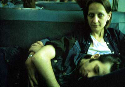 Two people sit with a dark blue seat back behind them. One, on the right of the image, looks to the camera with the other lays asleep on their lap across the bottom of the image.