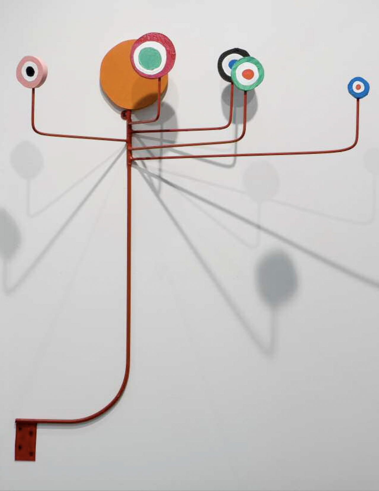 A sculpture affixed to the wall made of a red steel rod with red steel rod arms that project out from the top to make shadows against the wall.