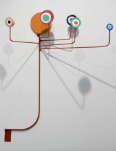 A sculpture affixed to the wall made of a red steel rod with red steel rod arms that project out from the top to make shadows against the wall.