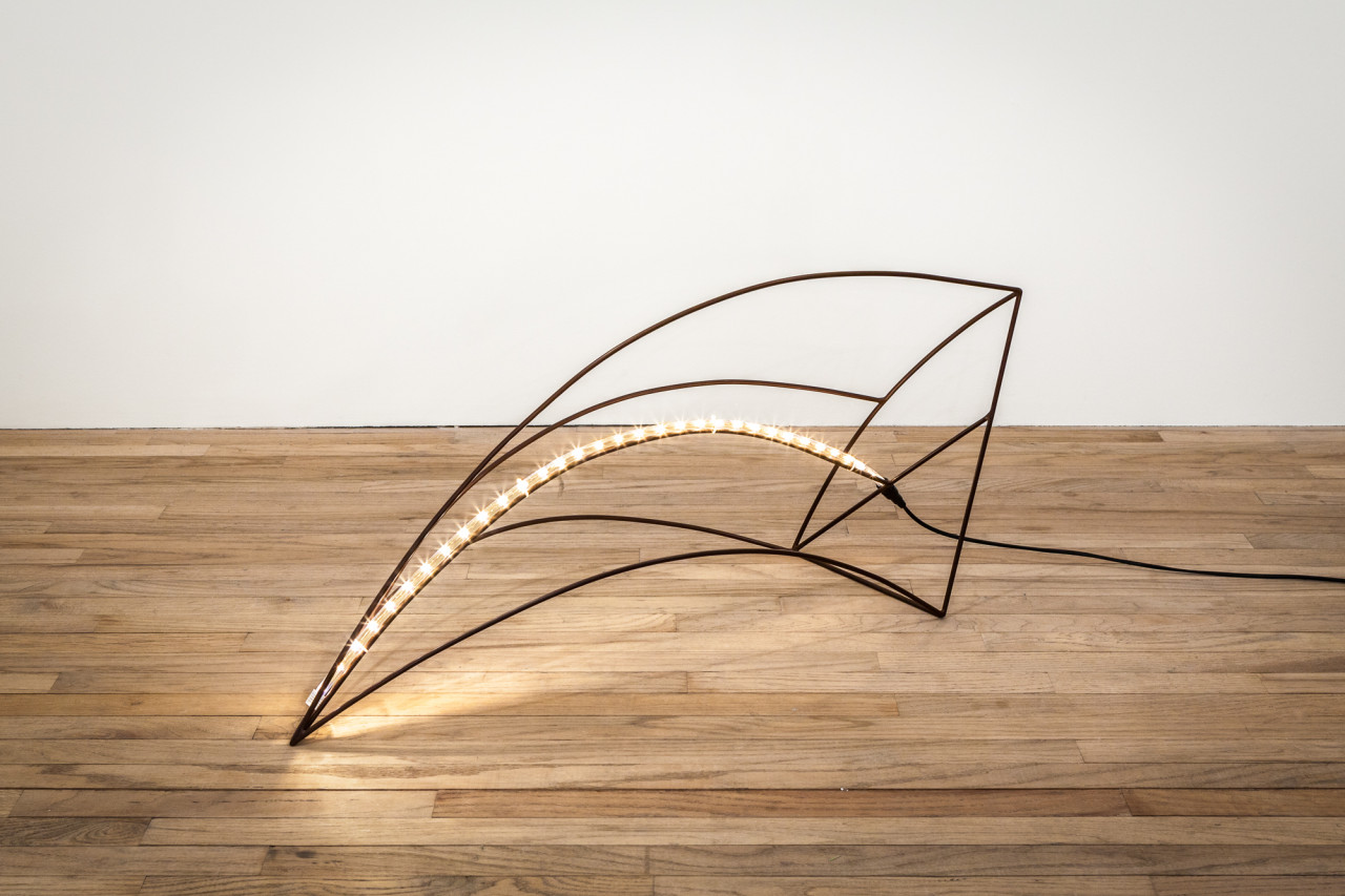 The photo shows steel rods that make a curved pyramid shape laying on its side with an LED light-strip running along one of the rods. The sculpture lies on a wooden floor against a white wall.