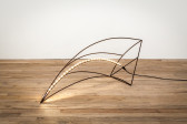 The photo shows steel rods that make a curved pyramid shape laying on its side with an LED light-strip running along one of the rods. The sculpture lies on a wooden floor against a white wall.