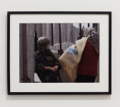 A photo in a black frame shows a person in military uniform, leaning against a concrete wall looking at a person who faces away from the camera with a patterned headscarf, red sweater, and light-yellow blanket.