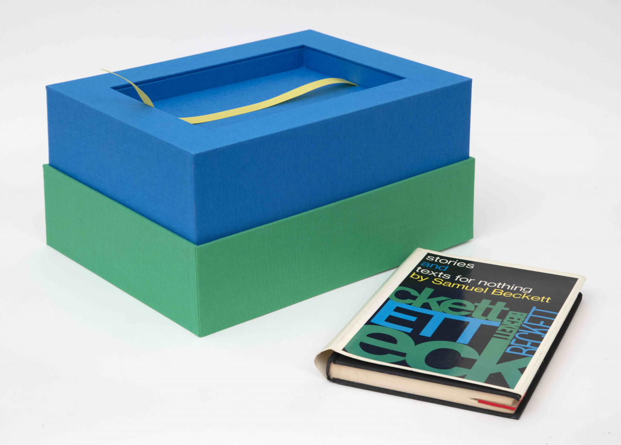A rectangular open box sits next to the hardcover book Stories and texts for nothing by Samuel Beckett, both against a white background. The green box lies open, exposing a blue interior with a rectangular cutout for the book to be placed in. A yellow ribbon functions as a pull-tab.
