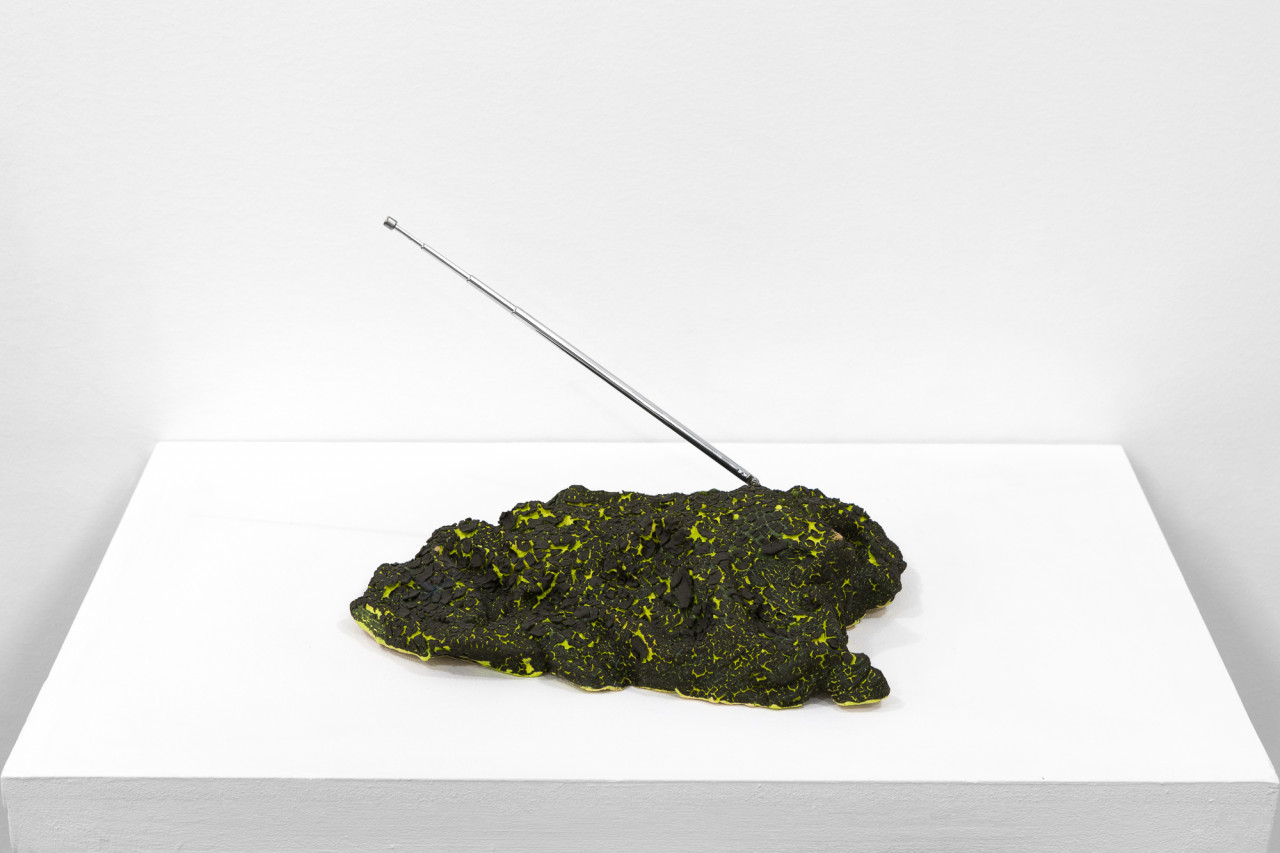 A green object with a cracked, black surface sits on a white base. A metal antenna extends out on a diagonal.